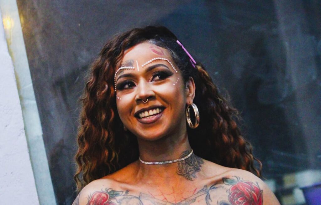 Foto de uma mulher negra de pele clara, possui cabelos longos cacheados da cor castanho claro, com uma presilha rosa do lado direito, tem uma tatuagem em formato de beijo na testa do lado direito e do lado esquerdo uma tatuagem de coroa em cima da sobrancelha, tem pedrinhas contornando suas duas sobrancelhas, no olha leva uma maquiagem preta, tem um piercing no septo, um brinco de argola dourado do lado direito e do lado esquerdo seu cabelo tampa a orelha, no pescoço tem uma gargantilha de pedras brilhantes,.