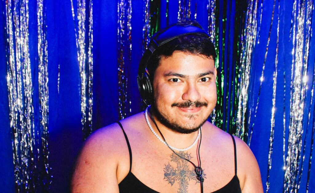 pessoa gender fluid, branca, está com headphone na cabeça, seu cabelo é liso, curto e preto, tem bigode, está usando um colar de pérolas e tem uma tatuagem de um crucifico no peito, usa uma blusa de alça fina. O fundo da imagem é uma cortina cintilante da cor azul.