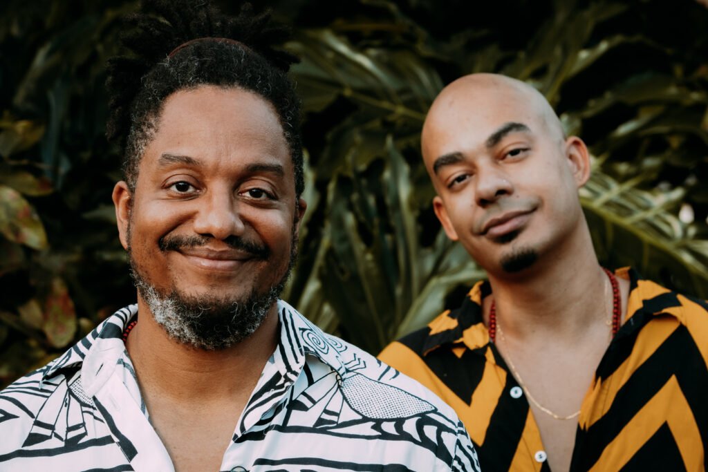 foto de dois homens negros em pé sorrindo para câmera. A esquerda o homem veste camisa social da cor branca com detalhes cinzas em forma de figuras geométricas, com os dois primeiro botões desabotoados. O cabelo está amarrado com um elástico da cor marrom, que por sua vez tem dreads e é grisalho. Possui bigode e barba também, grisalhas. Olha diretamente para a câmera e sorri. O homem do lado direito é careca, veste um blusa social amarela com formas geométricas da cor preta, com os dois primeiros botões, desabotoados. Tem cavanhaque ralo. Ao fundo tem folhas de arvores verdes desfocadas da foto.