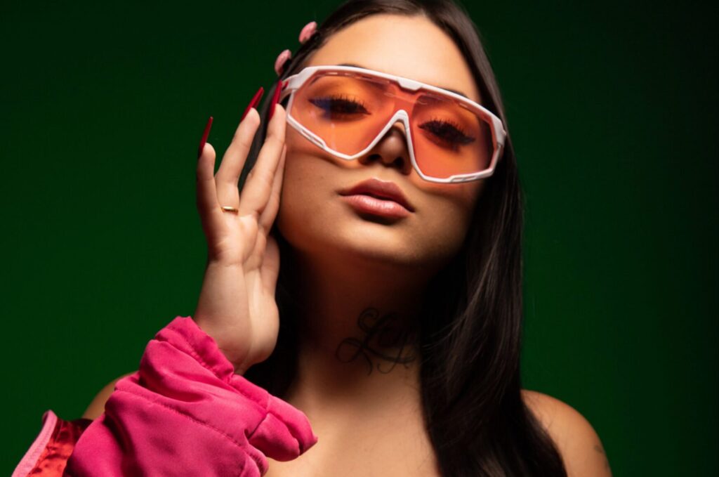 foto de uma mulher branca, aparecendo parte do seu caso rosa na mão esquerda que está levantada segurando o óculos que vai em seu rosto, com as unhas pintadas de vermelho. Seu cabelo está solto e, é liso, grande, da cor preta, com duas presilhas rosa do lado esquerdo, usa um óculos com armação rosa e lente alaranjada. Fundo da foto é degrade de verde escuro.