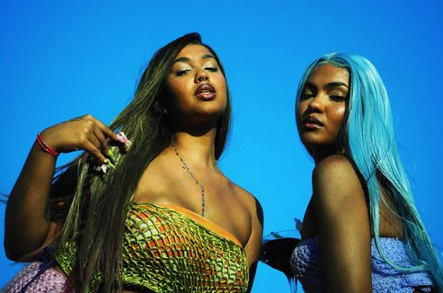 foto de duas mulheres negras, uma ao lado da outra a mulher da esquerda tem cabelos longos lisos e preto e está segurando com a mão esquerda, usa top verde, a mulher da esquerda tem o cableo liso da cor azul claro e está usando top azul. O fundo da foto é azul escuro.