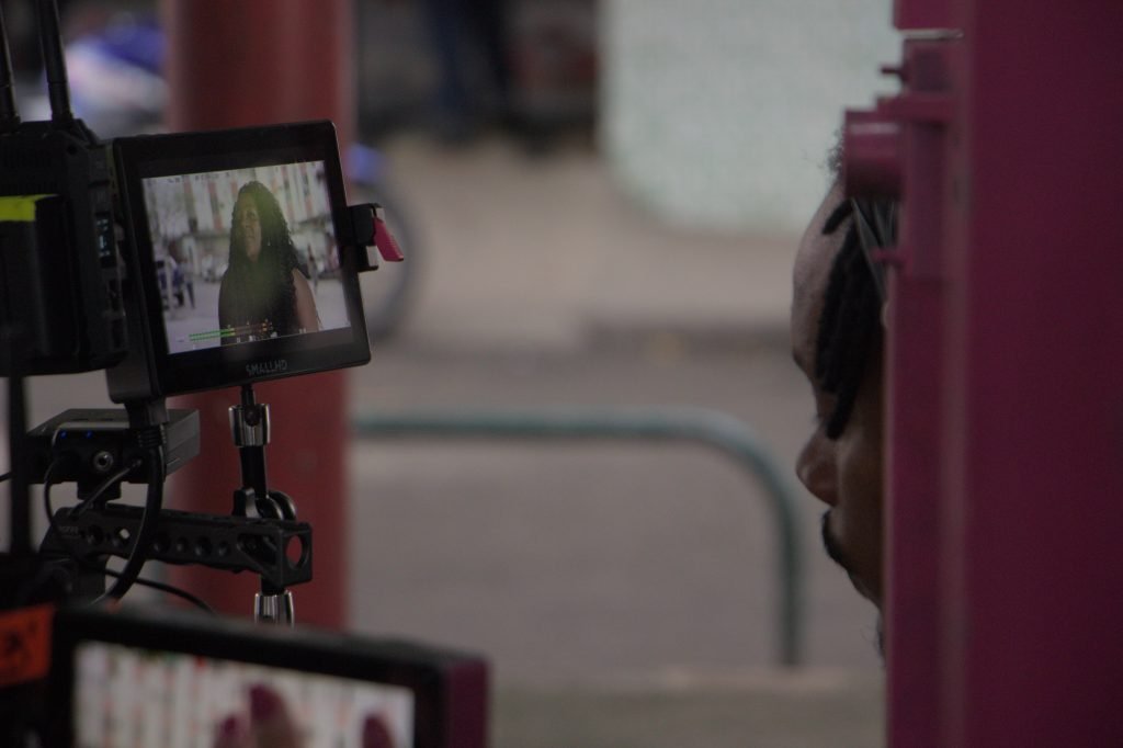 Foto colorida do visor de uma câmera de vídeo onde aparece uma mulher negra de cabelos compridos, marrom, encaracolados.